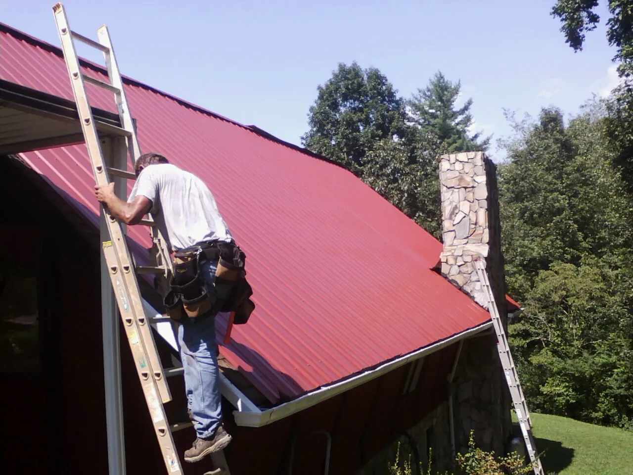 Roofer on a ladder beside a red metal roof and stone chimney — Soffit & Fascia Repair by Roofing Brothers in Portage